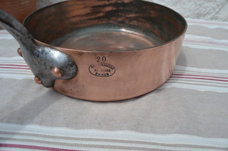 May include: A round copper saut&eacute; pan with a dark metal handle attached by three copper rivets. The side of the pan features the stamped text 20 and an oval mark reading Grands Magasins du Louvre Paris. The interior shows a metallic finish.