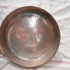 May include: A round copper frying pan with a metallic finish and visible circular scratch marks on the interior surface. The pan features a long, sturdy metal handle attached to the side with rivets, showing signs of wear and oxidation.