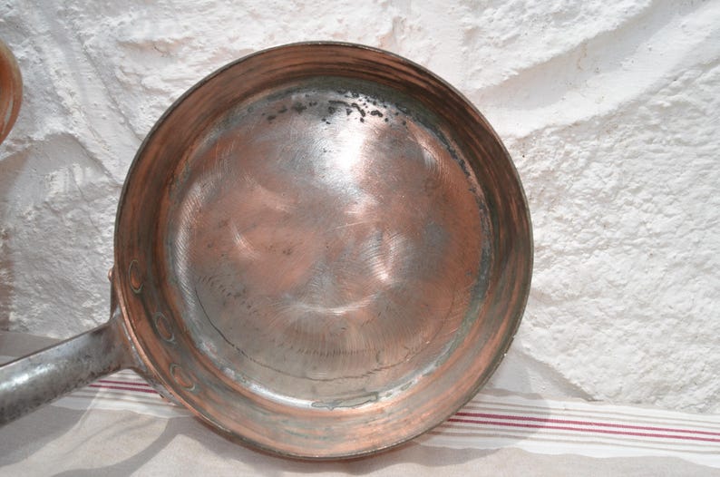 May include: A round copper frying pan with a metallic finish and visible circular scratch marks on the interior surface. The pan features a long, sturdy metal handle attached to the side with rivets, showing signs of wear and oxidation.