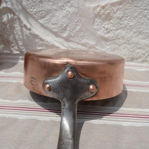May include: A round copper saut&eacute; pan featuring a sturdy iron handle attached with three copper rivets. The side of the pan displays a circular maker's mark stamp. The metal surface shows a warm, metallic finish with visible signs of use.