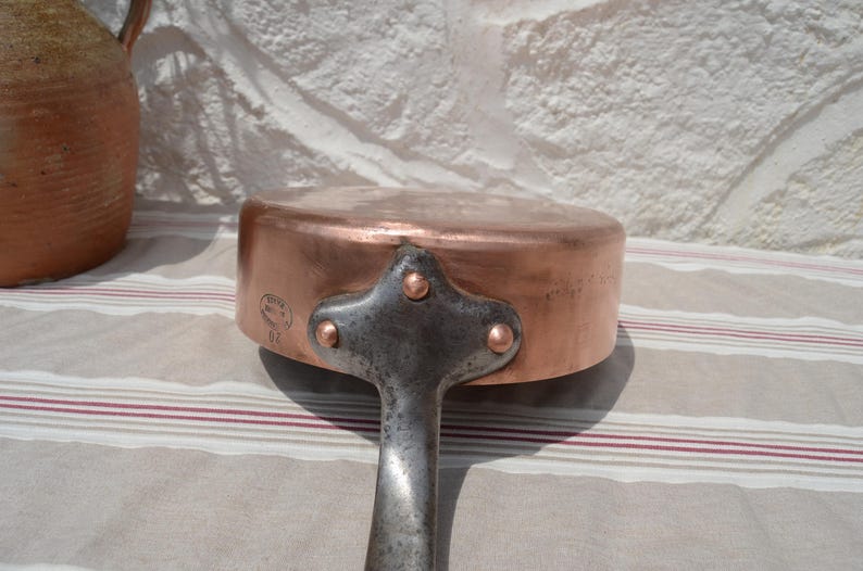 May include: A round copper saut&eacute; pan featuring a sturdy iron handle attached with three copper rivets. The side of the pan displays a circular maker's mark stamp. The metal surface shows a warm, metallic finish with visible signs of use.