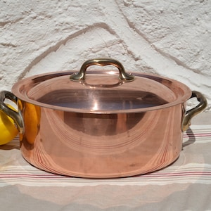 May include: A copper cooking pot with a lid and brass handles. The pot sits on a striped surface next to two yellow lemons. The pot has a shiny, reflective surface.