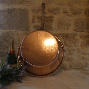 May include: A large copper pot with a wooden handle. The pot is sitting on a white tablecloth. There is a bottle of champagne and a bouquet of lavender flowers in the foreground.