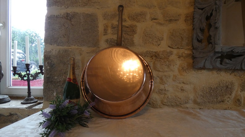 May include: A large copper pot with a wooden handle. The pot is sitting on a white tablecloth. There is a bottle of champagne and a bouquet of lavender flowers in the foreground.