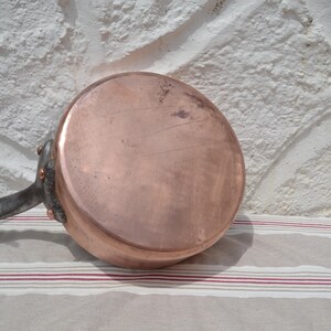 May include: A round copper saucepan with a flat base and a long, dark metal handle attached with three visible copper rivets. The exterior surface of the copper pan shows a polished finish with minor surface scratches and natural color variations.