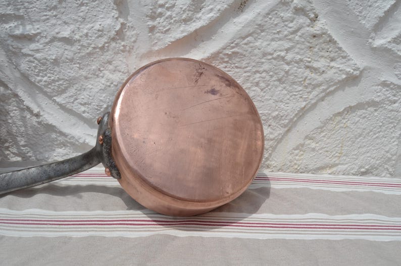 May include: A round copper saucepan with a flat base and a long, dark metal handle attached with three visible copper rivets. The exterior surface of the copper pan shows a polished finish with minor surface scratches and natural color variations.
