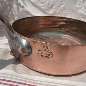 May include: A round copper saut&eacute; pan with a long, dark metal handle attached by two rivets. The side of the pan features the stamped text 20, LES MAGASINS DU LOUVRE, and PARIS within an oval border. The interior shows signs of wear and oxidation.