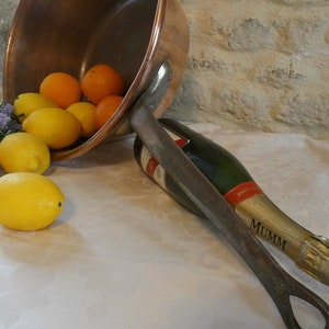 May include: A copper bowl filled with lemons and oranges, tipped over on a white tablecloth. A bottle of Mumm champagne is lying on its side next to the bowl, with a metal bottle opener.