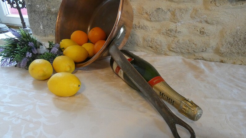 May include: A copper bowl filled with lemons and oranges, tipped over on a white tablecloth. A bottle of Mumm champagne is lying on its side next to the bowl, with a metal bottle opener.