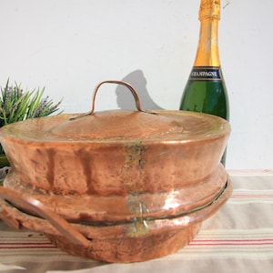 Könnte beinhalten: Ein großer Kupfertopf mit Deckel und zwei Griffen. Der Topf steht auf einer weißen Tischdecke mit roten Streifen. Im Hintergrund steht eine Flasche Champagner.