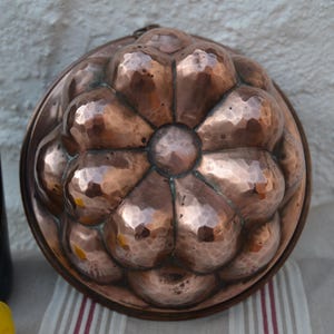 May include: A hammered copper mold with a floral design. The antique kitchenware is round with a central circle and petal-shaped indentations. A bottle of wine and a lemon are partially visible in the background.