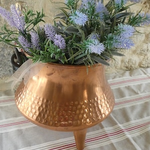 Peut inclure: Un pot en cuivre à trois pieds avec une texture martelée, rempli de fausses fleurs de couleur lavande et vertes. Le pot est posé sur une nappe à rayures, avec un mur en pierre en arrière-plan.