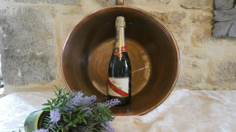 May include: A copper bowl with a bottle of champagne inside. The bowl is sitting on a white tablecloth. There is a bouquet of lavender flowers in the foreground.
