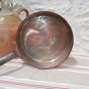 May include: A round copper pan with a long, dark metal handle. The interior of the pan shows a metallic surface with visible signs of wear and use. The pan is positioned next to a large, textured, tan and cream-colored ceramic jug.