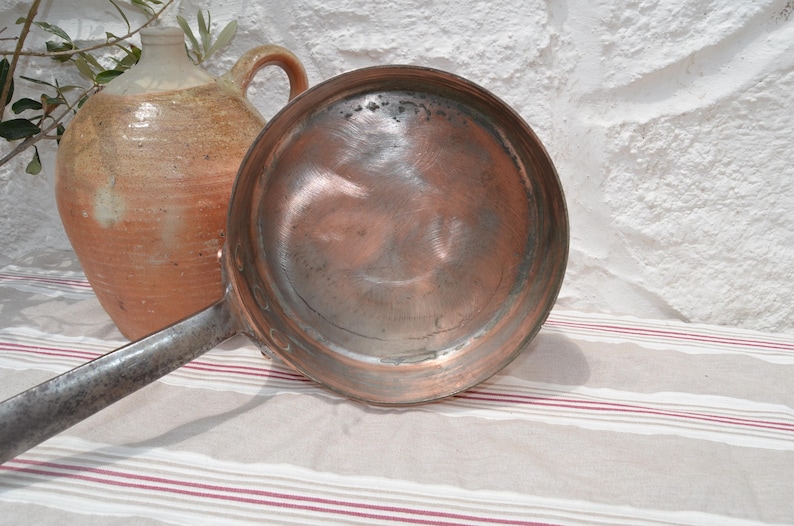 May include: A round copper pan with a long, dark metal handle. The interior of the pan shows a metallic surface with visible signs of wear and use. The pan is positioned next to a large, textured, tan and cream-colored ceramic jug.