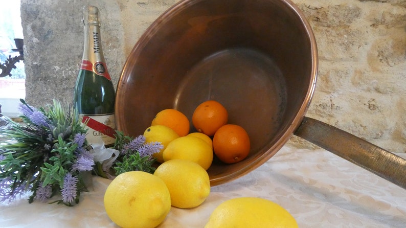May include: A copper pot with a long handle, filled with lemons and oranges. A bottle of champagne is in the foreground, next to a bouquet of lavender.