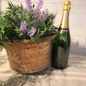 Pode incluir: Um balde de metal marrom rústico cheio de flores de lavanda roxas e folhagem verde. Uma garrafa de champanhe fica ao lado do balde sobre uma toalha de mesa listrada.