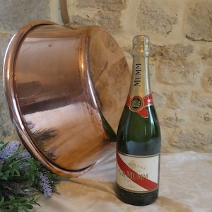 May include: A large copper pot with a hammered finish sits on a white tablecloth next to a bottle of Mumm champagne. The bottle has a red and white label with the text "Champagne G.H. Mumm Cordon Rouge".