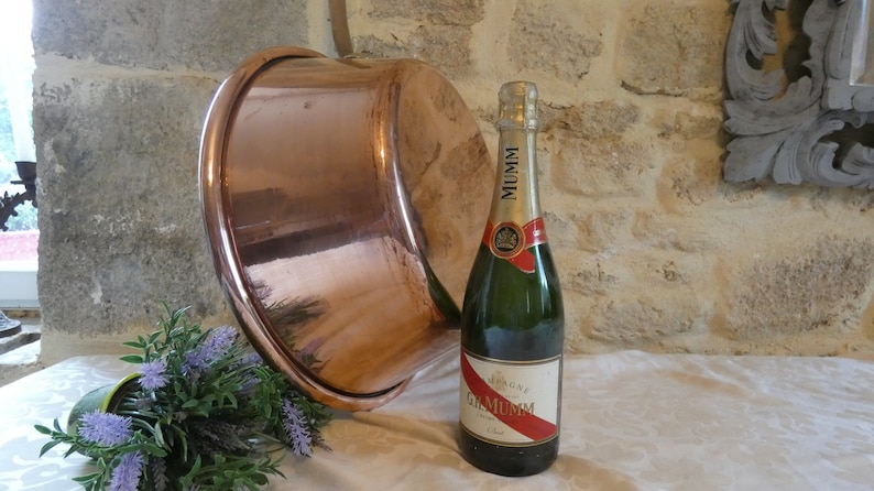 May include: A large copper pot with a hammered finish sits on a white tablecloth next to a bottle of Mumm champagne. The bottle has a red and white label with the text "Champagne G.H. Mumm Cordon Rouge".