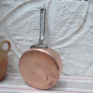 May include: A round copper pan with a long, dark metal handle attached by three copper rivets. The pan has a polished, metallic finish and a flat base. The handle features a small hole at the top for hanging, and the pan is positioned upright.