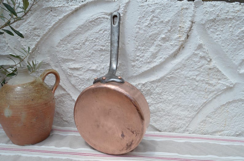 May include: A round copper pan with a long, dark metal handle attached by three copper rivets. The pan has a polished, metallic finish and a flat base. The handle features a small hole at the top for hanging, and the pan is positioned upright.