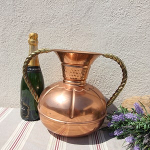 May include: A large copper vase with two twisted brass handles. The vase has a round body and a flared top. The vase is sitting on a striped tablecloth.