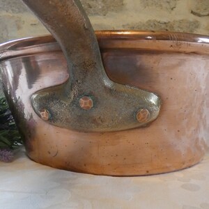 May include: A large copper pot with a heavy-duty handle. The pot has a rounded bottom and a wide, shallow opening. The handle is made of a darker metal and has four decorative rivets.