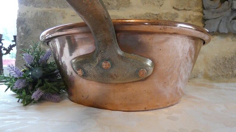 May include: A large copper pot with a heavy-duty handle. The pot has a rounded bottom and a wide, shallow opening. The handle is made of a darker metal and has four decorative rivets.