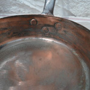 May include: A round copper frying pan with a metallic interior surface. The pan features a dark metal handle attached to the side with three visible circular rivets. The exterior walls show a warm copper tone with signs of oxidation and wear.