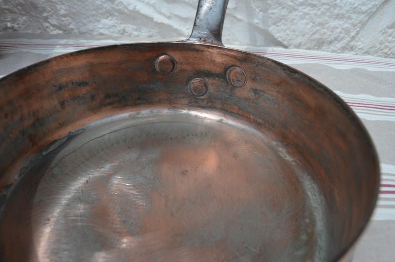 May include: A round copper frying pan with a metallic interior surface. The pan features a dark metal handle attached to the side with three visible circular rivets. The exterior walls show a warm copper tone with signs of oxidation and wear.