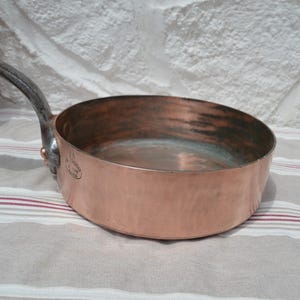May include: A round copper saut&eacute; pan with a long, dark, curved metal handle attached by a copper rivet. The exterior surface is polished copper, while the interior shows signs of use. A small oval stamp is visible on the side of the pan.