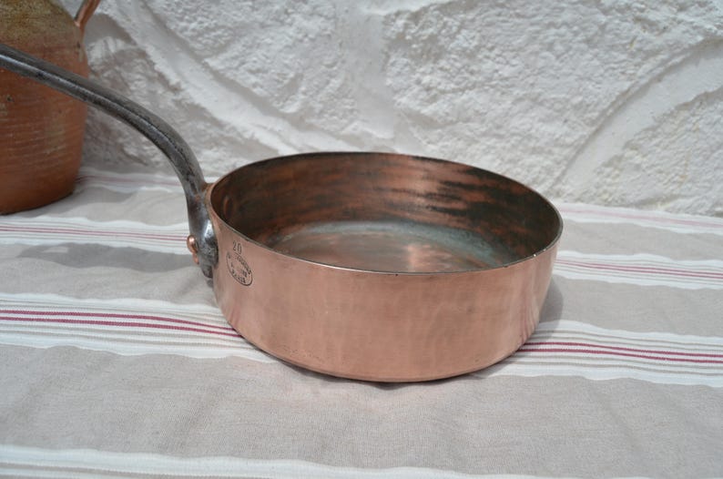 May include: A round copper saut&eacute; pan with a long, dark, curved metal handle attached by a copper rivet. The exterior surface is polished copper, while the interior shows signs of use. A small oval stamp is visible on the side of the pan.