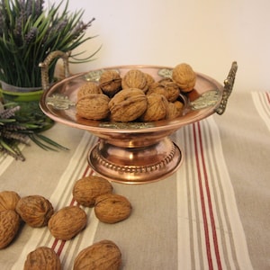 Peut inclure: Un bol de service en cuivre avec des poignées ornées et un piédestal, rempli de noix. Le bol est sur une nappe rayée.