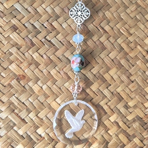 May include: A decorative hanging ornament featuring a clear, faceted circle with a dove design. The ornament includes a series of beads in various colors, including purple, clear, and blue, and a floral bead. The ornament is suspended by a silver chain.