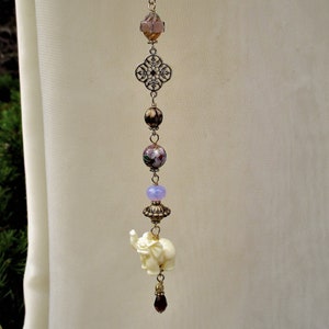 May include: A beaded charm featuring a cream-colored elephant pendant. The charm includes various beads in shades of purple, gold, and pink, along with a decorative filigree element. The charm is suspended from a black cord.