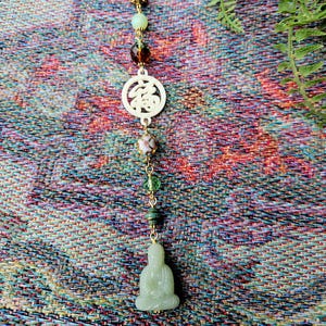 May include: A handcrafted pendant featuring a small jade Buddha figure, adorned with colorful beads and a white circular charm with an Asian symbol. The pendant hangs from a brown cord, showcasing a blend of natural and cultural elements.