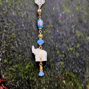 May include: A handcrafted beaded charm featuring an ivory-colored elephant pendant. The charm includes blue, gold, and floral beads, along with a silver filigree accent. The charm is suspended from a gold-colored chain.