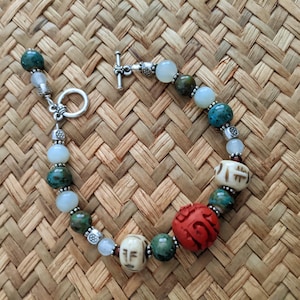 May include: A beaded bracelet featuring a variety of colorful beads. The bracelet includes turquoise, green, white, and red beads, with carved and patterned elements. The bracelet is set against a woven background.
