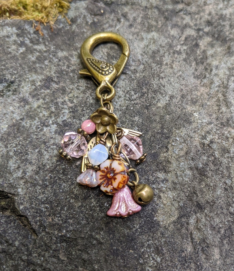 Pu&ograve; includere: Un ciondolo decorativo con una chiusura color ottone e un gruppo di perline e ciondoli. Il ciondolo presenta perline rosa, trasparenti e blu, insieme a una piccola campana e ciondoli a forma di fiore. Il design generale ha un'estetica vintage.