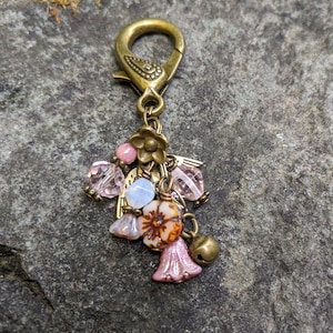 Pu&ograve; includere: Un ciondolo decorativo con un fermaglio color ottone e un gruppo di perline e ciondoli. Il ciondolo presenta perline rosa e trasparenti, piccoli fiori e una campana. Il design generale ha un'estetica vintage e artigianale.