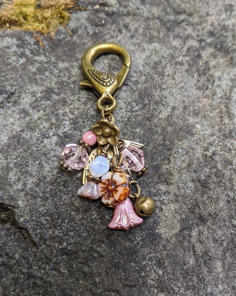 Pu&ograve; includere: Un ciondolo decorativo con un fermaglio color ottone e un gruppo di perline e ciondoli. Il ciondolo presenta perline rosa e trasparenti, piccoli fiori e una campana. Il design generale ha un'estetica vintage e artigianale.