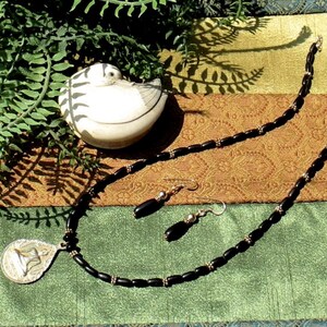 May include: A black beaded necklace and matching earrings set with a silver pendant. The pendant has a unique design. A small, white ceramic bird figurine with black stripes is also in the image. The jewelry is displayed on a patterned fabric.