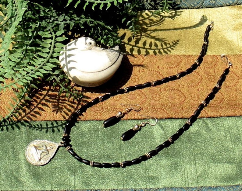 May include: A black beaded necklace and matching earrings set with a silver pendant. The pendant has a unique design. A small, white ceramic bird figurine with black stripes is also in the image. The jewelry is displayed on a patterned fabric.