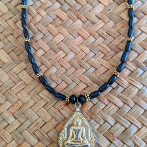May include: A black beaded necklace with a gold-colored pendant. The pendant features a seated Buddha figure. The necklace is displayed on a woven, light brown surface.