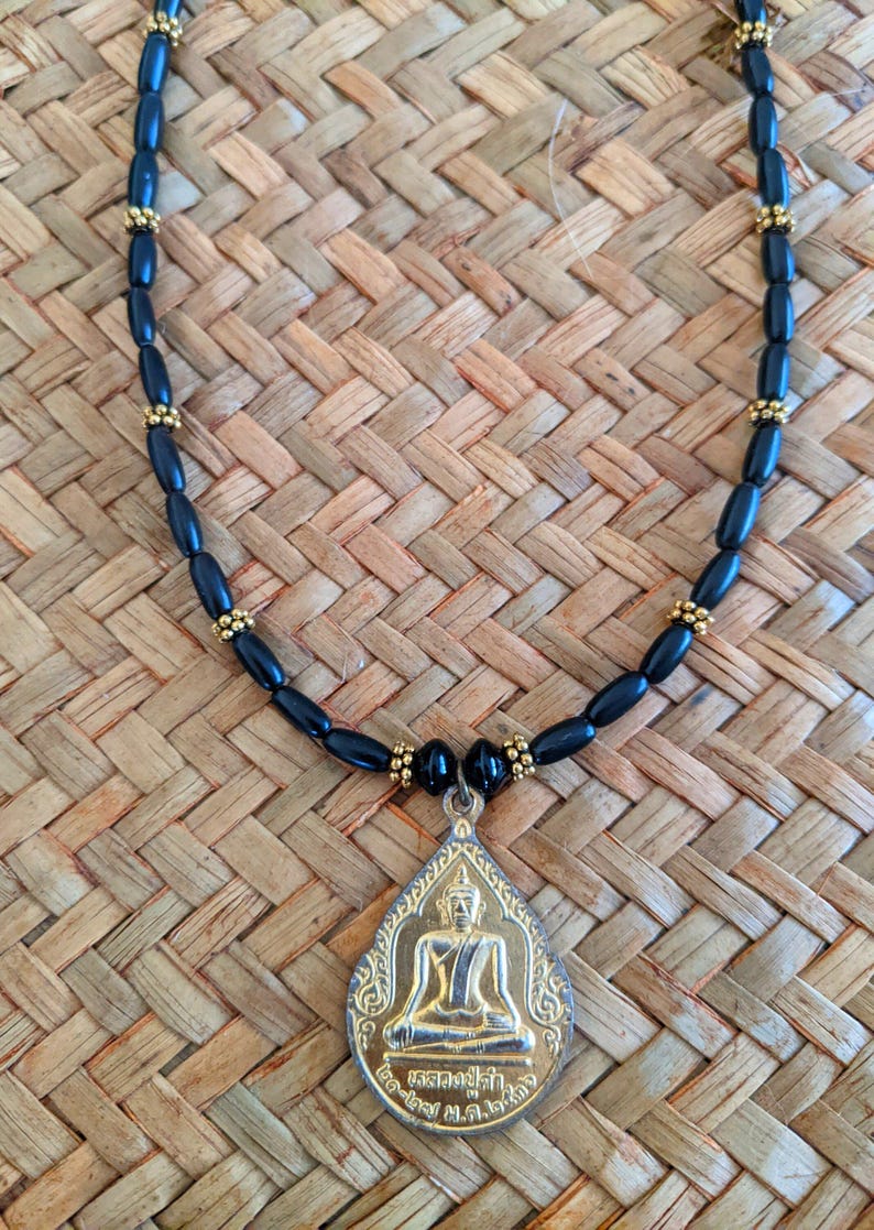May include: A black beaded necklace with a gold-colored pendant. The pendant features a seated Buddha figure. The necklace is displayed on a woven, light brown surface.