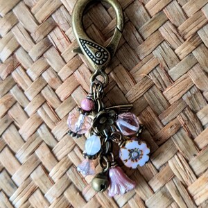 Pu&ograve; includere: Un fermaglio decorativo color bronzo con un motivo floreale e un gruppo di ciondoli. I ciondoli includono perline rosa e trasparenti, una piccola campana e perline a forma di fiore rosa. Il fermaglio &egrave; posto su uno sfondo intrecciato.