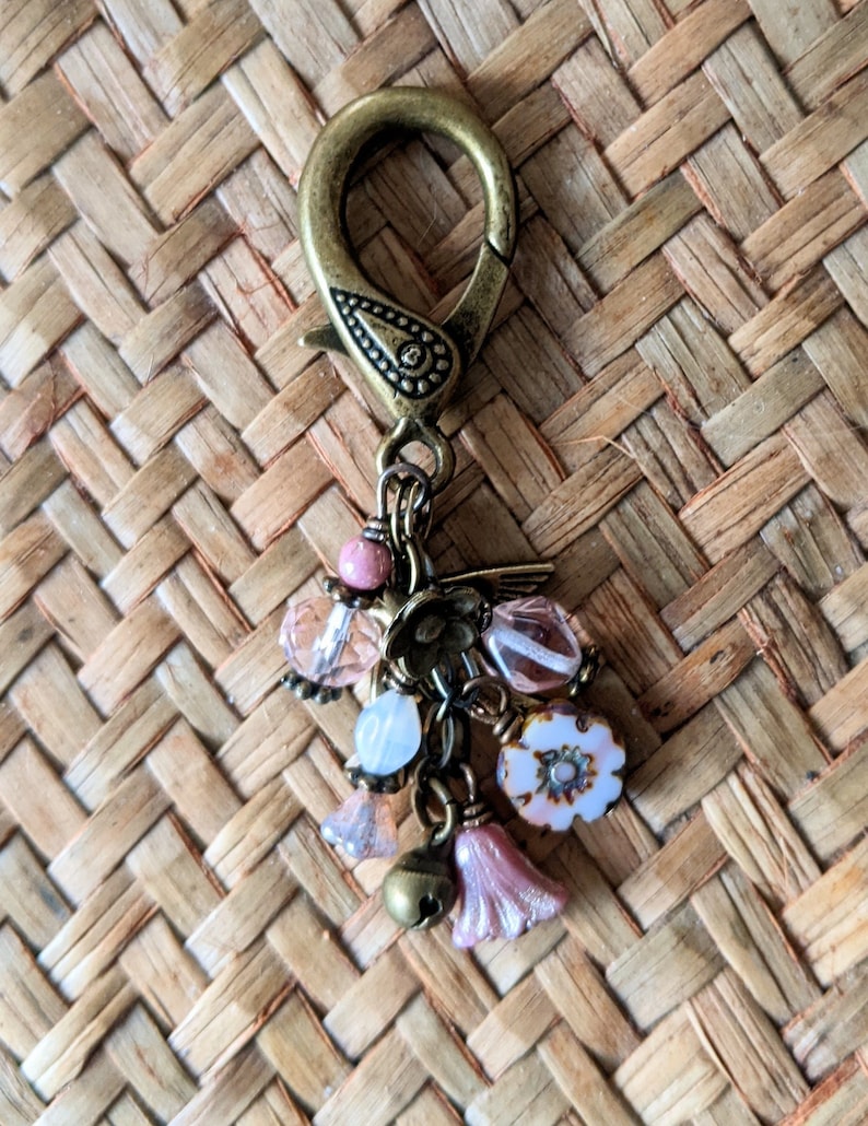Pu&ograve; includere: Un fermaglio decorativo color bronzo con un motivo floreale e un gruppo di ciondoli. I ciondoli includono perline rosa e trasparenti, una piccola campana e perline a forma di fiore rosa. Il fermaglio &egrave; posto su uno sfondo intrecciato.