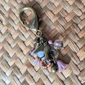 Pu&ograve; includere: Un portachiavi decorativo con chiusura e ciondolo color bronzo. Il ciondolo presenta un uccellino, perline rosa e trasparenti, campanellini e dettagli a forma di fiore. Lo sfondo &egrave; un materiale intrecciato marrone chiaro.