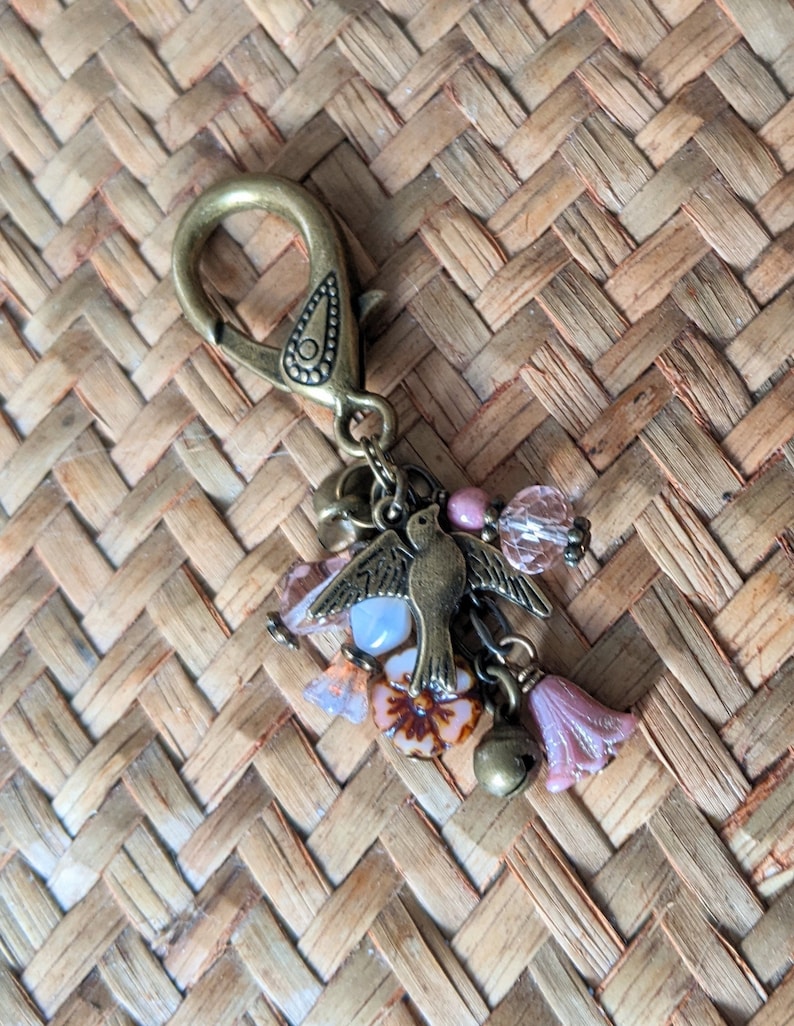 Pu&ograve; includere: Un portachiavi decorativo con chiusura e ciondolo color bronzo. Il ciondolo presenta un uccellino, perline rosa e trasparenti, campanellini e dettagli a forma di fiore. Lo sfondo &egrave; un materiale intrecciato marrone chiaro.