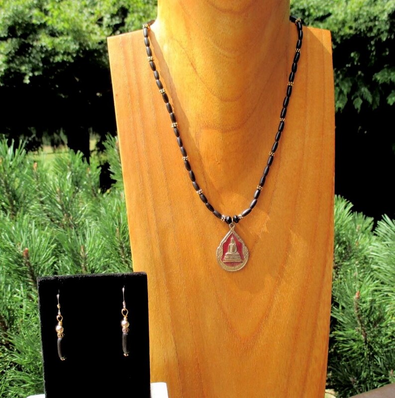 May include: A necklace and earrings set featuring a pendant with a Buddha image. The necklace has black beads with gold-colored accents. The earrings have a similar design. The pendant has a red background.